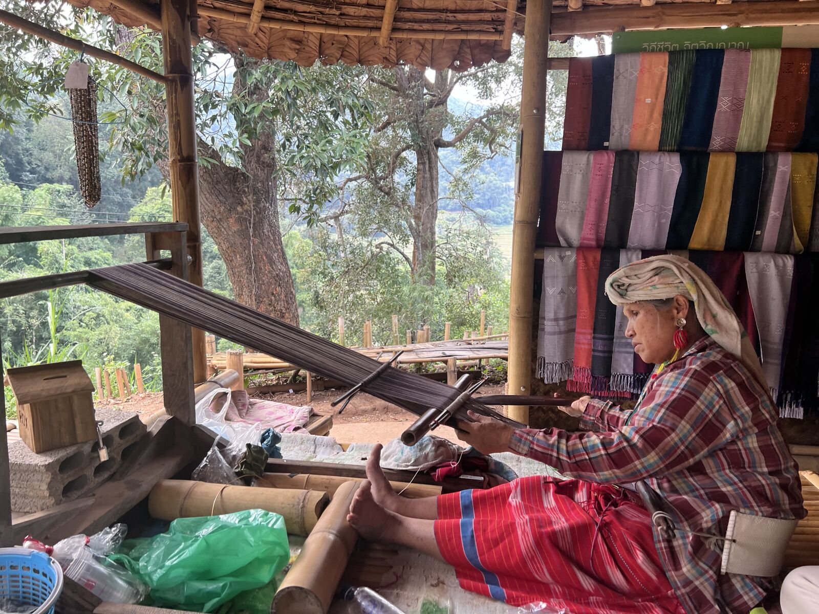 A local hill-tribe woman at her loom with fabric, forest, and open air in Chiang Mai.
